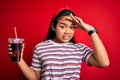 Young asian girl drinking cola fizzy refreshment using straw over isolated red background stressed with hand on head, shocked with Royalty Free Stock Photo