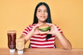 Young asian girl eating a tasty classic burger smiling looking to the side and staring away thinking
