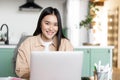 Young asian girl freelancer working remotely from home. Woman with laptop studying indoors from her kitchen, smiling at Royalty Free Stock Photo