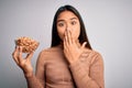 Young asian girl holding bowl of healthy peanuts eating as snack for diet over white background cover mouth with hand shocked with Royalty Free Stock Photo