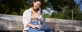 Young asian girl talking on mobile phone, using laptop, working online from park, sitting on bench on sunny day with Royalty Free Stock Photo