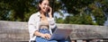 Young asian girl talking on mobile phone, using laptop, working online from park, sitting on bench on sunny day with Royalty Free Stock Photo