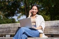 Young asian girl talking on mobile phone, using laptop, working online from park, sitting on bench on sunny day with Royalty Free Stock Photo