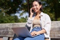 Young asian girl talking on mobile phone, using laptop, working online from park, sitting on bench on sunny day with Royalty Free Stock Photo