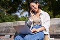 Young asian girl talking on mobile phone, using laptop, working online from park, sitting on bench on sunny day with Royalty Free Stock Photo