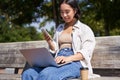 Young asian girl talking on mobile phone, using laptop, working online from park, sitting on bench on sunny day with Royalty Free Stock Photo