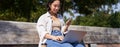 Young asian girl talking on mobile phone, using laptop, working online from park, sitting on bench on sunny day with Royalty Free Stock Photo