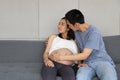Young asian husband in blue T shirt comforting, embracing his young unhappy asian pregnant belly wife while sitting on sofa in