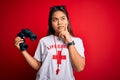 Young asian lifeguard girl wearing t-shirt with red cross using whistle and binoculars serious face thinking about question, very