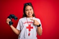 Young asian lifeguard girl wearing t-shirt with red cross using whistle and binoculars very happy pointing with hand and finger