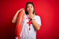 Young asian lifeguard girl wearing t-shirt with red cross using whistle holding orange float cover mouth with hand shocked with