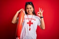 Young asian lifeguard girl wearing t-shirt with red cross using whistle holding orange float doing ok sign with fingers, excellent