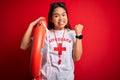 Young asian lifeguard girl wearing t-shirt with red cross using whistle holding orange float screaming proud and celebrating