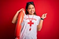 Young asian lifeguard girl wearing t-shirt with red cross using whistle holding orange float very happy pointing with hand and