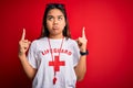 Young asian lifeguard girl wearing t-shirt with red cross using whistle over isolated background Pointing up looking sad and