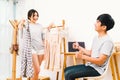 Young Asian lover couple choosing cloth together at home. Girlfriend holding 2 dress, boyfriend thumbs up.
