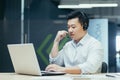 A young Asian male consultant. Sitting in the office, wearing headphones, talking from a laptop Royalty Free Stock Photo