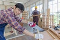 Young asian male worker wearing plaid shirt and blue jean apron with white gloves holding saw cutting wood stick on the table full Royalty Free Stock Photo