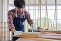 Young asian male worker wearing plaid shirt and blue jean apron with white gloves holding saw cutting wood stick on the table full Royalty Free Stock Photo