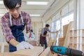 Young asian male worker wearing plaid shirt and blue jean apron with white gloves holding saw cutting wood stick on the table full Royalty Free Stock Photo