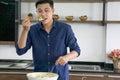 Young asian man in blue shirt stands in kitchen scoops green vegetable into his mount. Cooking salad Diet healthy