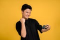 Young Asian man in a cheerful black T-shirt is taking a selfie with a mobile phone showing the winner`s gesture. Yellow background Royalty Free Stock Photo