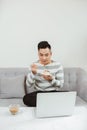 Young Asian man eating cake when sitting on sofa and using laptop Royalty Free Stock Photo