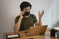 Young man in face mask talking on mobile pone and using laptop computer at cafe.