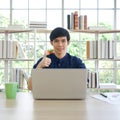 Young asian man lifts his thumb up with a smile behind laptop computer in the library. There are coffee mug, computer, pen, and