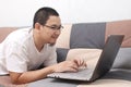 Young Asian man lying on sofa working remotely from home, using laptop computer having video call or buying shopping online via Royalty Free Stock Photo