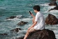 Young Asian man with mobile smart phone sitting on the rock of natural seashore. Internet of things concept. Royalty Free Stock Photo