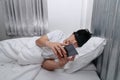 Young Asian man with smartphone lying down on bed in bedroom. Nomophobia symptoms or no mobile phone phobia concept. Royalty Free Stock Photo