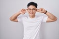 Young asian man standing over white background doing peace symbol with fingers over face, smiling cheerful showing victory Royalty Free Stock Photo