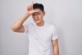 Young asian man standing over white background making fun of people with fingers on forehead doing loser gesture mocking and