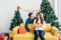 Young Asian man surprises her girlfriend that wearing Santa Claus hat with a Christmas gift at home with a Christmas tree.
