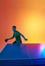 Young Asian man, table tennis athlete preparing for return, hit ball in neon light against warm yellow gradient