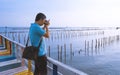 Young Asian man taking picture with digital camera on the old multicolored wooden bridge at sea viewpoint Royalty Free Stock Photo