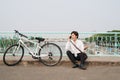 Young asian man talking by mobile phone while sitting on the ground near the bike, outdoors. Wearing stylish clothes Royalty Free Stock Photo