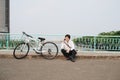 Young asian man talking by mobile phone while sitting on the ground near the bike, outdoors. Wearing stylish clothes. Royalty Free Stock Photo