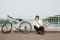 Young asian man talking by mobile phone while sitting on the ground near the bike, outdoors. Wearing stylish clothes. Royalty Free Stock Photo