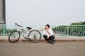 Young asian man talking by mobile phone while sitting on the ground near the bike, outdoors. Wearing stylish clothes Royalty Free Stock Photo