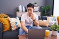 Young asian man using laptop at home sitting on the sofa pointing to the eye watching you gesture, suspicious expression Royalty Free Stock Photo