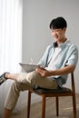 Young Asian man using tablet, designing his artwork on tablet, sitting on a chair in living room Royalty Free Stock Photo