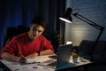 Young Asian man wearing a red T-shirt is writing papers on a desk in the dark late at night at home Royalty Free Stock Photo