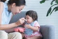 Young Asian mother holding spoon feeding child, Caucasian Cute 7 month newborn baby girl eating blend mashed food, sitting on sofa Royalty Free Stock Photo