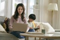 young asian mother working on laptop while son playing computer game using digital tablet at home Royalty Free Stock Photo