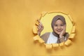 Young Asian muslim teenager smiling poses through torn yellow paper hole pointing a copy space or blank yellow paper, wearing Royalty Free Stock Photo