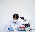 Young Asian scientist in latex gloves and medical mask writing in notebook near test tubes and microscope Royalty Free Stock Photo