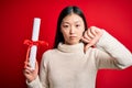 Young asian student woman holding graduate diploma over red  background with angry face, negative sign showing dislike Royalty Free Stock Photo