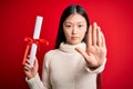 Young asian student woman holding graduate diploma over red isolated background with open hand doing stop sign with serious and Royalty Free Stock Photo